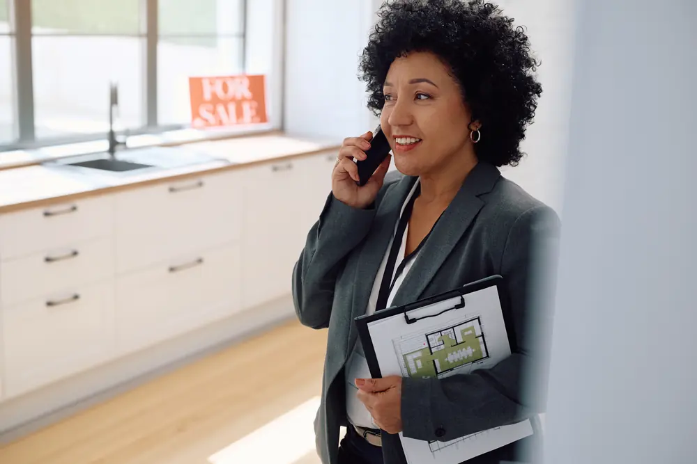 Real-estate agent on the phone in a kitchen holding a floor plan, with a for-sale sign behind her